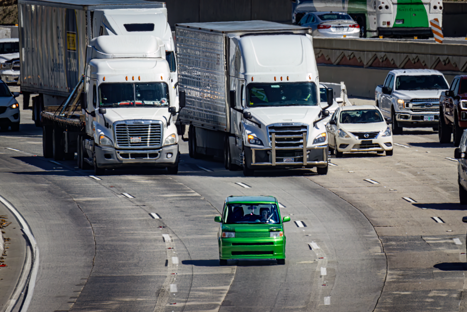 A small bright green car driving on a multi-lane road surrounded by various larger vehicles, including white box trucks, a white sedan, and other cars, with clear daylight and urban traffic environmen