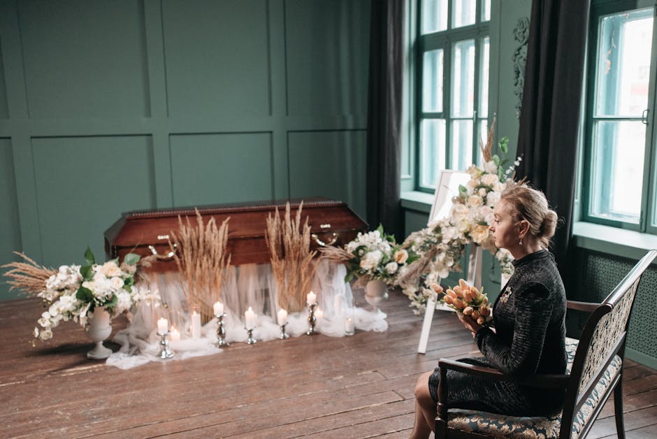 A woman seated in a black patterned dress holds a small bouquet of peach and cream flowers as she faces an elegant floral arrangement. The arrangement features white roses, peonies, and greenery place